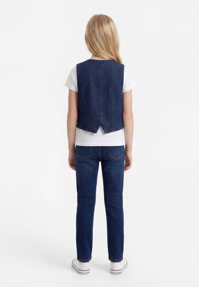 Hosszú szőke hajú gyermek háttal áll, fehér pólót, sötétkék farmer mellényt, farmert és fehér tornacipőt visel, egyszínű fehér háttér előtt.