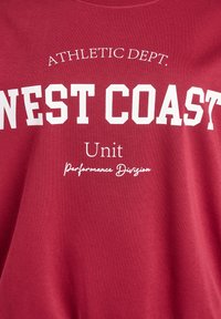 Maron farvet bomulds T-shirt med fed hvid tekst: "ATHLETIC DEPT. WEST COAST" med yderligere mindre tekst, der siger "Unit Performance Division."