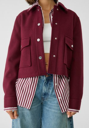 Personne portant une veste bordeaux boutonnée par-dessus une chemise rayée rouge et blanche, un crop top blanc et un jean bleu, coupée au niveau de la taille.
