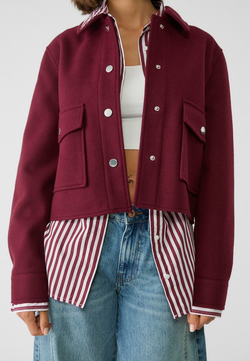 Personne portant une veste bordeaux boutonnée par-dessus une chemise rayée rouge et blanche, un crop top blanc et un jean bleu, coupée au niveau de la taille.