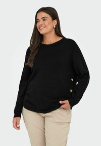 Donna sorridente con lunghi capelli scuri, che indossa un maglione nero con bottoni laterali e pantaloni beige, in piedi con una mano in tasca.