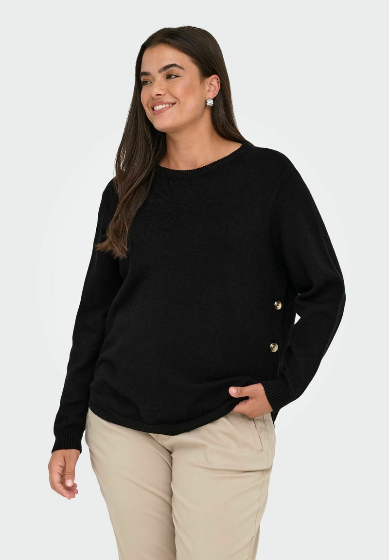 Donna sorridente con lunghi capelli scuri, che indossa un maglione nero con bottoni laterali e pantaloni beige, in piedi con una mano in tasca.