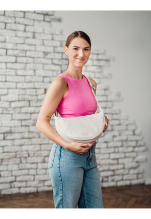 Weiße halbkreisförmige Tasche aus weichem Material, mit einem einzelnen Schultergurt und einem dezenten blauen Logo, getragen von einem Model in Jeans.