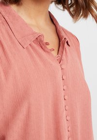 Blouse rose texturée avec un col, comprenant des détails de boutons et une patte de boutonnage. Le tissu est léger avec un subtil effet côtelé et des boucles décoratives.