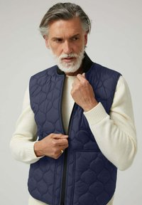 Chaleco acolchado azul marino con un cuello alto, cierre de cremallera y bolsillo delantero, usado sobre un suéter crema. Tejido texturizado con patrón de cuadrícula acolchada.