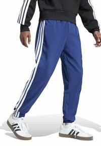 Pantalones deportivos azules con rayas blancas en los lados, cinturilla elástica, piernas ajustadas y un material ligero. Combinados con zapatillas blancas.