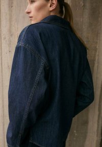 Veste en denim bleu foncé à coupe décontractée, avec un col pointu, des épaules tombantes et des détails de couture minimalistes.