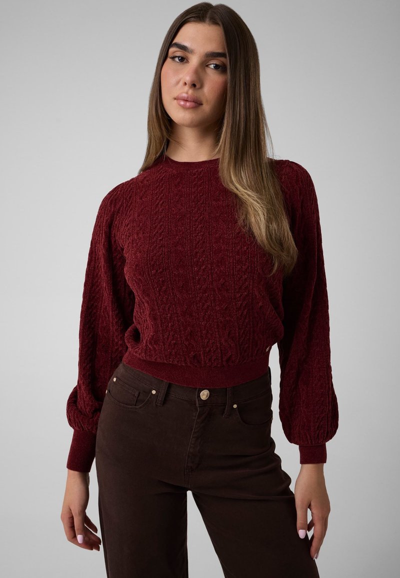 Jeune femme aux longs cheveux raides portant un pull texturé bordeaux et un pantalon marron foncé sur un fond clair uni.