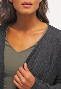 Žena se střední barvou pleti, která má na sobě tmavě šedý svetr s výstřihem do V přes olivově zelené tričko, drží svetr u hrudi.