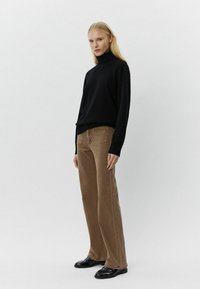 Maglione a collo alto nero con maniche lunghe, abbinato a pantaloni larghi marrone chiaro e mocassini neri, in piedi su uno sfondo neutro.