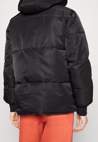 Giacca a vento nera con design trapuntato, dotata di cappuccio, polsini elasticizzati e una superficie liscia simile alla satinata. Indossata con pantaloni arancioni.