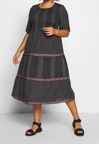 Vestido midi negro con mangas cortas, que presenta un patrón texturizado de pequeños puntos blancos. El dobladillo está adornado con un colorido borde bordado. Sandalias negras.
