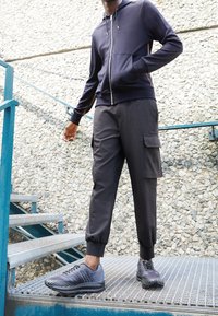 Sweat à capuche zippé bleu marine en tissu doux, associé à un pantalon cargo noir avec poches latérales, et des chaussures de sport foncées à semelle texturée.
