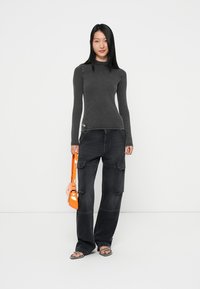 Grijze blouse met lange mouwen, hoge hals, strakke snit. Zwarte cargobroek met grote zakken. Oranje handtas, platte sandalen. Eenvoudige achtergrond.