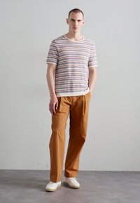 Chemise en tricot à rayures à manches courtes couleur crème, avec des lignes multicolores. Associée à un pantalon beige et des baskets blanches, sur un fond neutre.