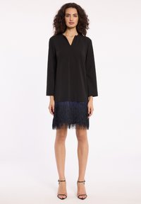 Schwarzes Kleid mit V-Ausschnitt, langen Ärmeln und einem fransigen blauen Saum. Der Stoff hat eine glatte Textur, die im Kontrast zu der Textur der Fransen steht.