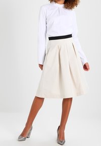 Jupe plissée beige avec une taille noire, associée à une chemise blanche à manches longues et des chaussures à talons hauts en argent métallisé. Texture lisse.