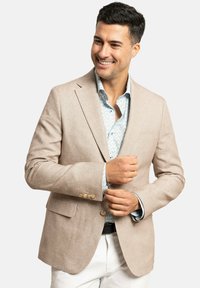 Blazer de lino beige con solapas de pico, dos bolsillos frontales y detalles en botones. Llevado sobre una camisa de patrón floral en azul claro.