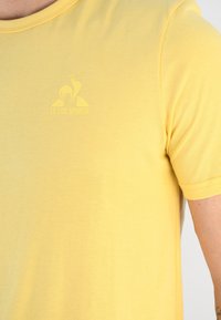 Camiseta amarilla de manga corta elaborada con tejido suave, que presenta un sutil logo impreso "LE COQ SPORTIF" en el pecho y un cuello redondo.