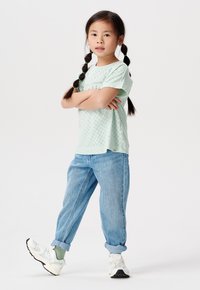 Camicetta verde chiaro a maniche corte con dettagli in pizzo, abbinata a jeans in denim blu arrotolati e sneakers bianche con accenti neri.