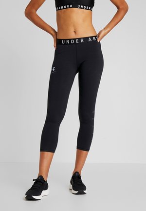 Kobieta w czarnych krótkich leginsach Under Armour i dopasowanym sportowym staniku, stojąca z rękami na biodrach na jednolitym tle.