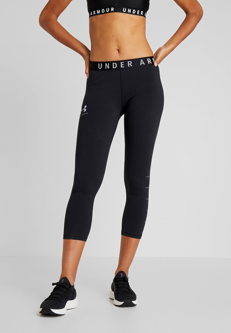 Femme portant un legging court noir Under Armour et un brassière de sport assortie, debout les mains sur les hanches sur un fond uni.
