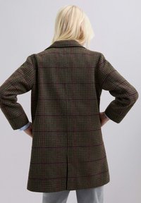 Persona rubia vestida con un abrigo de patrón de houndstooth marrón con líneas de cuadro púrpura, con las manos en las caderas, de espaldas frente a un fondo sencillo.