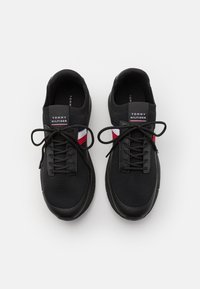 Baskets noires à lacets avec une tige en mesh, présentant un accent rayé rouge et blanc, et une semelle en caoutchouc noire. Logo sur la languette.