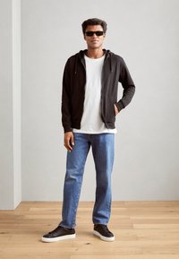 Schwarze Zip-Hoodie über einem weißen T-Shirt, kombiniert mit hellblauen Jeans und schwarzen Sneakers. Das Model trägt dunkle Sonnenbrillen. Schlicher Hintergrund.