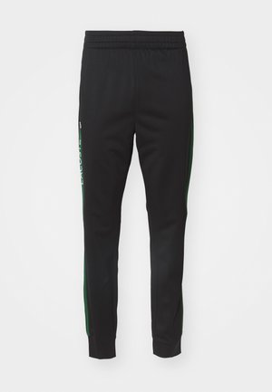 Pantalones deportivos negros hechos de una tela suave; cuentan con franjas laterales verdes y un logo blanco de Lacoste. Cintura elástica para mayor comodidad.