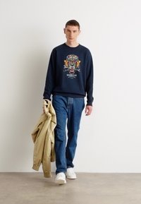 Navy sweatshirt with a tiger graphic, blue jeans, holding a beige jacket. Model wearing white sneakers, in a neutral backdrop.