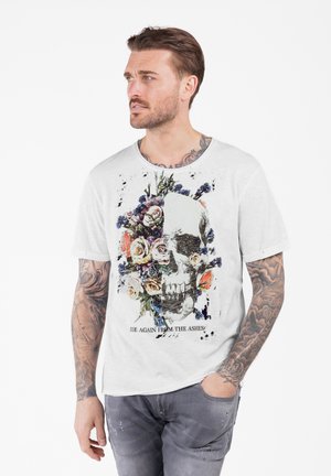 Uomo con tatuaggi che indossa una maglietta bianca con un design che presenta metà teschio e metà floreale e il testo "Ritorna dalle ceneri" mentre guarda di lato.