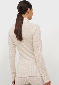 Aansluitend lang mouwtop in lichtbeige met een subtiel geometrisch patroon, textuurstof en een hoge hals. Heeft aansluitende mouwen en een zoom.