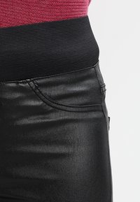 Svarta faux läderbyxor med en ribbad svart midja. Har en sidficka och en glansig yta som framhäver de texturerade detaljerna.