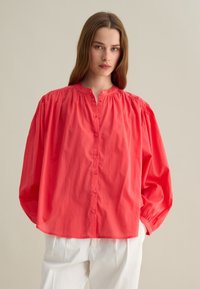 Camicia rosa corallo con maniche a sbuffo, colletto alto e dettagli arricciati sulle spalle. Abbinata a pantaloni ampi di colore chiaro.