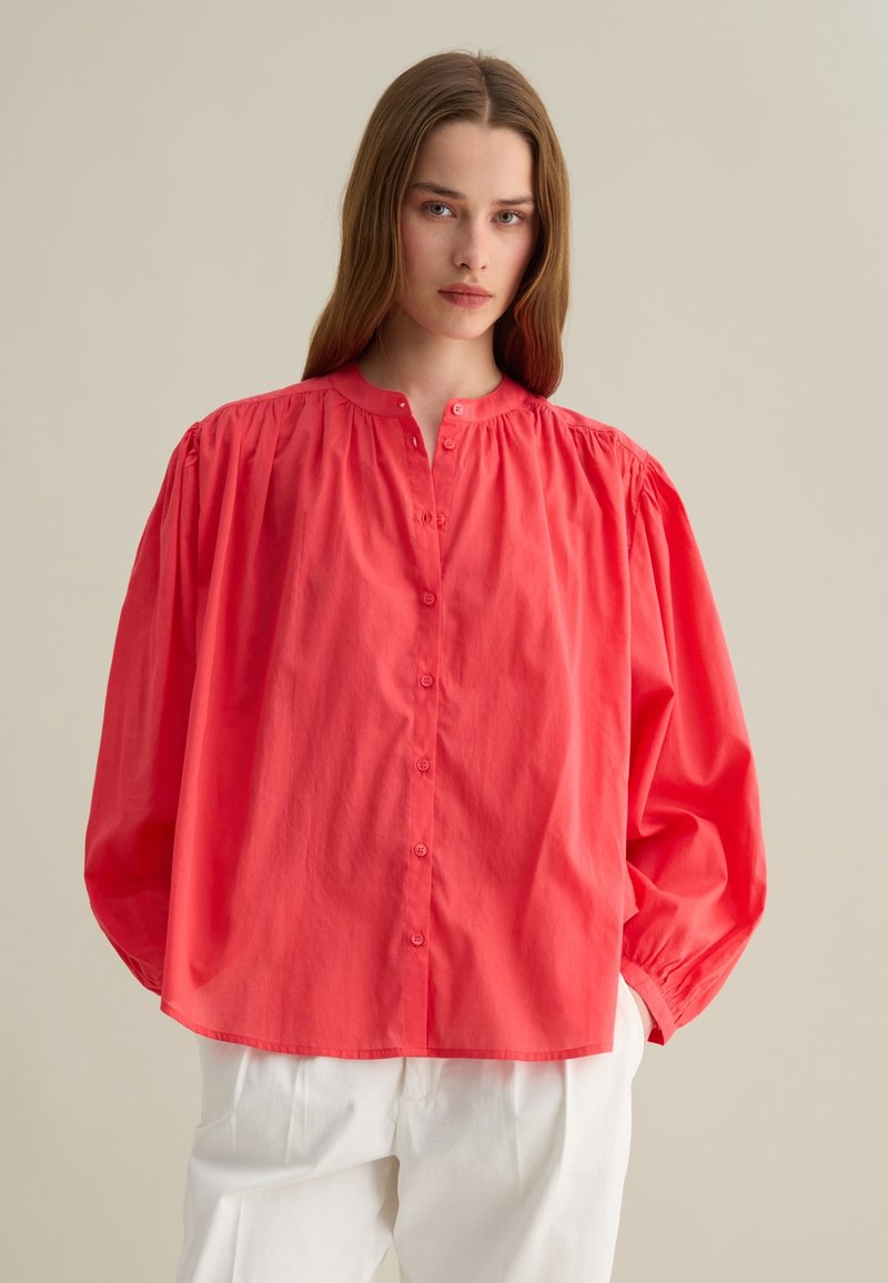 Camicia rosa corallo con maniche a sbuffo, colletto alto e dettagli arricciati sulle spalle. Abbinata a pantaloni ampi di colore chiaro.