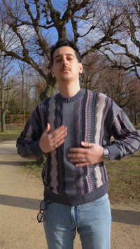 Άνδρας που φοράει πουλόβερ με σχέδια και τζιν στέκεται σε ένα μονοπάτι σε πάρκο με γυμνά δέντρα, αγγίζοντας το στήθος του με τα δύο χέρια.