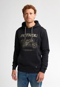 Uomo con capelli ricci che indossa un felpa nera con una grafica di motocicletta e testo "PETROL", abbinato a jeans blu, in piedi su uno sfondo bianco.