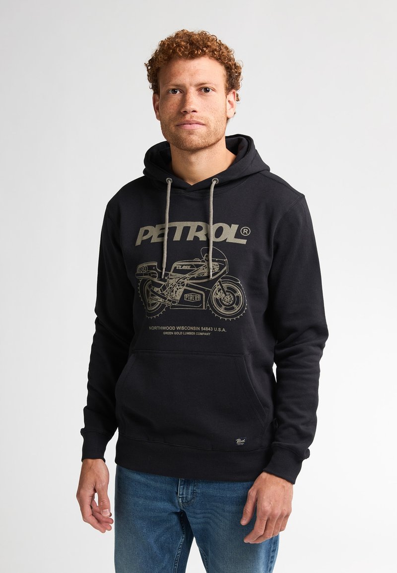 Uomo con capelli ricci che indossa un felpa nera con una grafica di motocicletta e testo "PETROL", abbinato a jeans blu, in piedi su uno sfondo bianco.