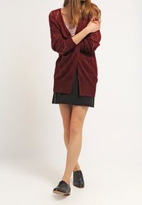 Vinröd stickad cardigan med lös passform, V-hals och diamantsmönster. Kombinerad med en svart kjol och svarta snörskor.