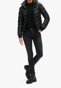 Persona che indossa un giubbotto imbottito nero, un maglione nero, jeans skinny grigio scuro, una cintura nera e stivali da combattimento neri con lacci, in piedi con una mano in tasca.