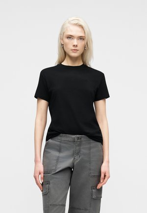 Blond kvinne står vendt forover iført svart kortermet skjorte og grå cargobukser mot en ensfarget lys bakgrunn.