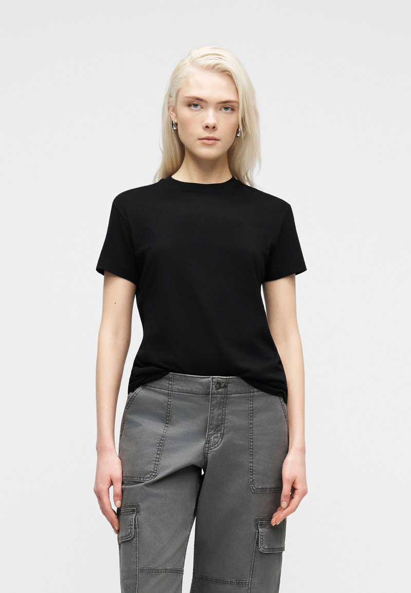 Ξανθιά γυναίκα στέκεται μπροστά, φοράει μαύρο κοντομάνικο πουκάμισο και γκρι παντελόνι cargo, πάνω σε απλό ανοιχτό φόντο.