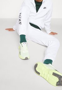 Vit zip-up träningsdräkt med svart "NIKE" text på ärmarna, parat med gröna strumpor och ljusgröna sneakers med svarta accenter och mönstrade sålar.