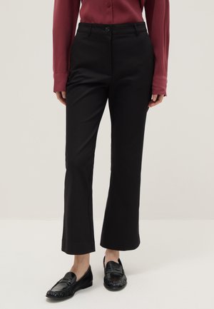 Vrouw die zwarte broek met rechte pijpen, zwarte loafers en een bordeauxrode overhemd met lange mouwen draagt, staand op een lichte achtergrond.