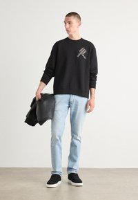 Hanorac negru cu detalii albe ale logo-ului, mâneci lungi, guler rotund. Asociat cu blugi albaștri deschiși și adidași negri, ținând o jachetă gri.