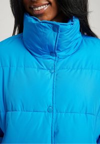 Chaqueta acolchada de color azul brillante con cuello alto, que presenta cierres de broche y textura acolchada. Tela suave y brillante con un diseño cálido e aislante.