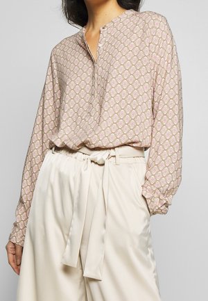 Femme portant un chemisier à manches longues à motifs, rentrée dans un pantalon taille haute couleur crème avec une ceinture en tissu nouée.