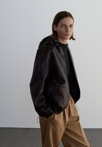 Giacca in pelle nera con cappuccio, vestibilità oversize, texture liscia. Abbinata a una camicia nera a pois e pantaloni beige a vita alta.