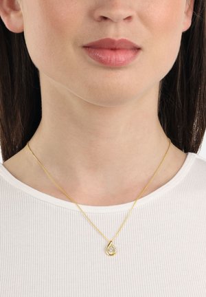 Collar de pendant de oro con forma de lágrima y acento de piedra clara, suspendido de una fina cadena de oro sobre una blusa blanca de canalé.